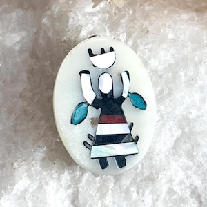 Puede incluir: Colgante ovalado con un diseño nativo americano. El colgante presenta un fondo blanco con una figura en negro, rojo y blanco, con acentos turquesa. La figura tiene una corona y los brazos extendidos.