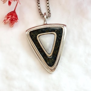 May include: A triangular silver pendant on a box chain necklace. The pendant features a dark, textured, moss-green inlay surrounding a smaller, smooth, white triangular stone set in the center. The metal frame is polished silver.