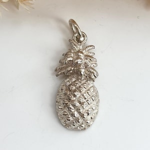 Puede incluir: Un dije de piña plateada con una superficie texturizada y un lazo en la parte superior para sujetarlo a un collar o pulsera. El dije es una representación en miniatura detallada de una fruta de piña.