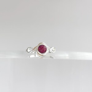 K&ouml;nnte beinhalten: Ein silberner Ring mit einem runden, tief rosa-roten Stein in der Mitte. Die Ringschiene ist auf beiden Seiten der zentralen Steinfassung mit einem Unendlichkeitssymbol gestaltet, wodurch ein durchgehendes Schleifenmuster entsteht.