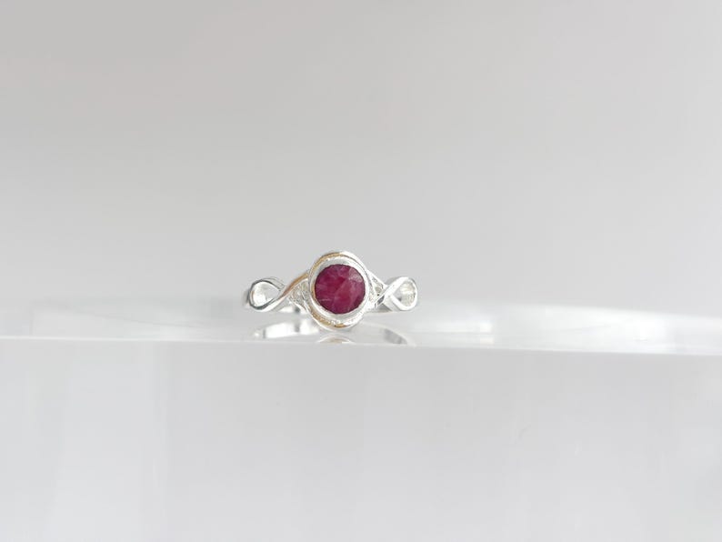K&ouml;nnte beinhalten: Ein silberner Ring mit einem runden, tief rosa-roten Stein in der Mitte. Die Ringschiene ist auf beiden Seiten der zentralen Steinfassung mit einem Unendlichkeitssymbol gestaltet, wodurch ein durchgehendes Schleifenmuster entsteht.
