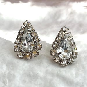 May include: A pair of vintage teardrop-shaped earrings. Each earring features a large, clear, pear-cut crystal surrounded by smaller, clear crystals. The earrings have a silver-tone metal setting and are displayed on a white, textured surface.