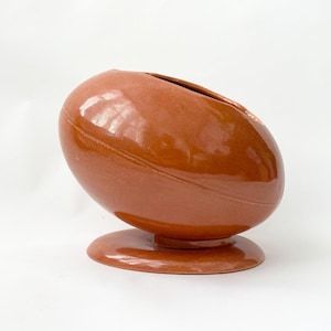 Op de afbeelding: Bruine keramische vaas in de vorm van een rugbybal, met een cirkelvormige basis. De vaas heeft een grote opening aan de bovenkant en gestikte details. De vaas is ongeveer 25 cm lang.