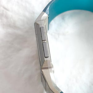 May include: A digital watch with a silver-toned rectangular casing and two rectangular side buttons. The watch features a vibrant turquoise blue silicone strap that curves around the side of the device, showcasing a sleek and functional design.