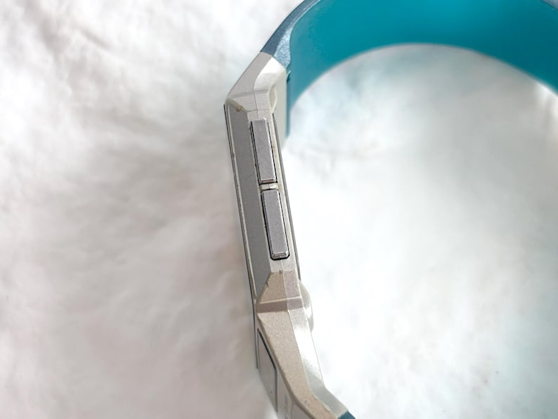 May include: A digital watch with a silver-toned rectangular casing and two rectangular side buttons. The watch features a vibrant turquoise blue silicone strap that curves around the side of the device, showcasing a sleek and functional design.