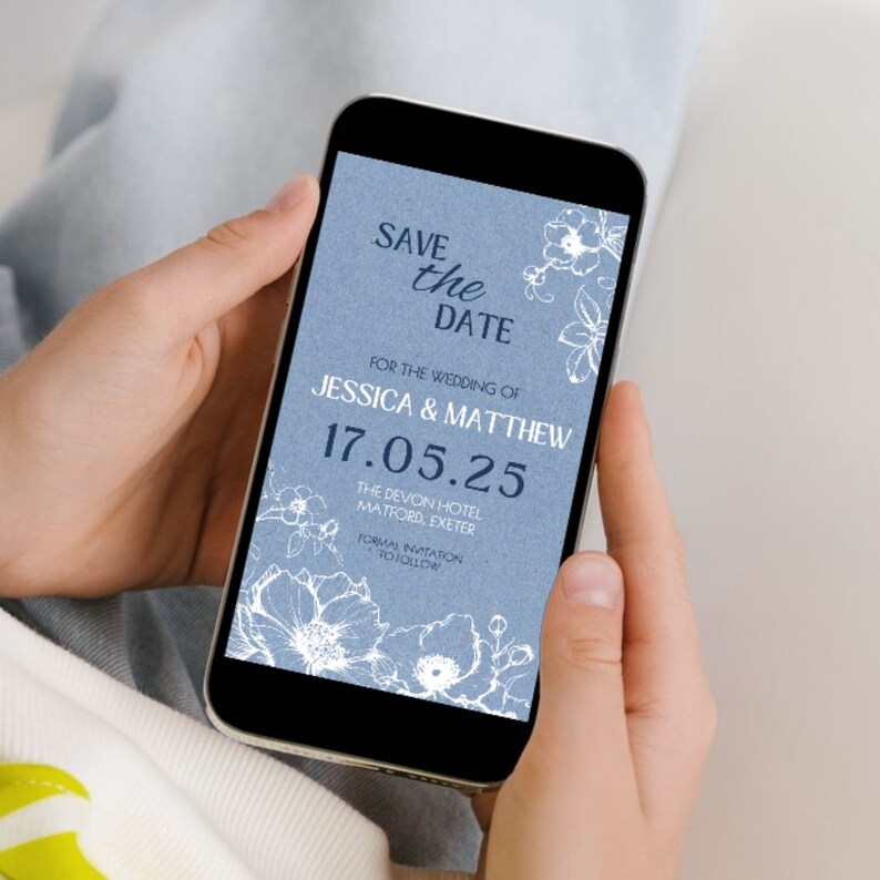 May include: A blue and white save the date card on a smartphone screen. The card features a floral design and the text "Save the Date" for the wedding of Jessica & Matthew on 17.05.25 at The Devon Hotel, Matford, Exeter. Formal invitation to follow.