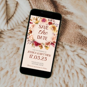 May include: A smartphone displays a wedding invitation with the text "SAVE the DATE." The design features a floral wreath in shades of peach, burgundy, and gold. The invitation includes the names Jessica & Matthew, the date 17.05.25, and the location.