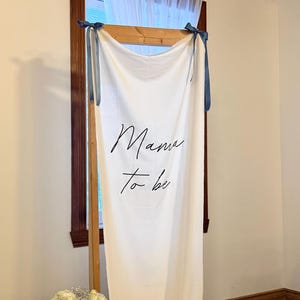 Può includere: Un banner di tessuto bianco con le parole "Mama to be" in scrittura nera è esposto su una cornice di legno. Nastri blu sono legati agli angoli superiori. Un vaso di rose bianche è a sinistra.