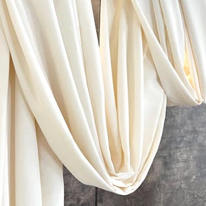 May include: Off-white fabric draped over a wooden rod, creating soft folds and shadows. The fabric hangs against a textured, dark gray wall. The overall aesthetic is minimalist and elegant.
