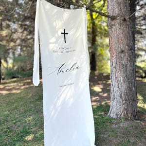 May include: A white fabric banner with a black cross and text, suspended from a tree branch. The banner reads "WELCOME TO THE CHRISTENING OF Amelia September 10, 2025". The fabric appears soft and flowing.