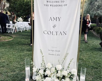Könnte beinhalten: Ein hohes, vertikales Hochzeits-Willkommensschild aus weißem Stoff, das an einem goldfarbenen Metallrahmen hängt. Der Text lautet: WELCOME TO THE WEDDING OF AMY & COLTAN, Las Vegas, Nevada, November 2, 2024. Weiße Rosen schmücken den Fuß des Schildes.