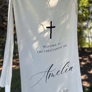 May include: Cream-coloured fabric banner featuring a black cross and the words "WELCOME TO THE CHRISTENING OF Amelia September 10, 2025." The banner is suspended from a tree branch, with the text in a simple, elegant font.