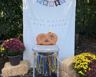 Może przedstawiać: Białe tło z tkaniny z banerem z napisem PROST oraz tekstem HAPPY BIRTHDAY DANE ANDREW. Pluszowy precel z małą żółtą czapeczką urodzinową znajduje się na krzesełku do karmienia ozdobionym niebieskimi, żółtymi i zielonymi wstążkami.