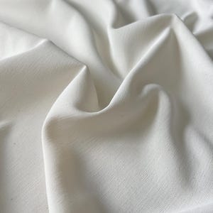 Può includere: Primo piano di un tessuto bianco con texture e pieghe visibili. Il tessuto appare di un colore bianco sporco uniforme, con sottili variazioni di tono dovute alla luce e alle ombre. Il materiale ha una leggera lucentezza.