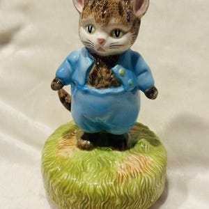 Puede incluir: Figura de cerámica de un gato con un atuendo azul, de pie sobre una base de hierba verde. El gato tiene pelaje marrón y canela, ojos verdes y nariz rosa. Un objeto decorativo.