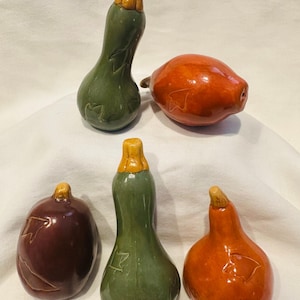 May include: A collection of ceramic gourds in various shapes and colors. The gourds are green, orange, and burgundy, with tan stems. Some gourds have leaf-like carvings on their surfaces. The arrangement suggests a fall harvest theme.
