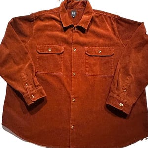 May include: A long-sleeve button-down shirt in a rust-coloured corduroy fabric. The shirt features a standard collar, a central button placket, and two chest pockets with buttoned flaps. The cuffs are also buttoned, and the brand label GAP is visible.