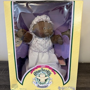 Puede incluir: Muñeca Cabbage Patch Kids Preemie vintage de 1985 en su caja original. La muñeca tiene piel oscura, un vestido blanco y un gorro. La caja es amarilla con un borde verde y presenta el logotipo de Cabbage Patch Kids y el logotipo de March of Dimes.
