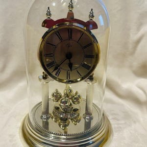 May include: A silver-toned clock with a gold-colored face, Roman numerals, and a glass dome. The clock has a pendulum and is displayed on a silver base. The clock is approximately 8 inches tall.