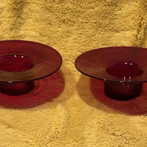 May include: Two matching, deep red glass bowls with a wide, shallow dish shape and a central, rounded base. The bowls are displayed on a yellow, textured surface, casting shadows.