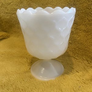 May include: A white, vintage-style glass bowl with a scalloped rim and a diamond pattern. The bowl sits on a small, round base. The bowl is likely used for serving or display.