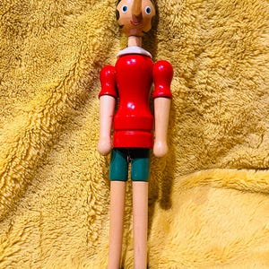 May include: Wooden Pinocchio doll with a red hat, shirt, and shorts. The doll has a long nose, blue eyes, and black shoes. The arms and legs are made of wood. The doll is standing on a yellow, textured surface.