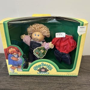 May include: A Cabbage Patch Kids Playmate doll in its original box. The doll has blonde hair, blue eyes, and is wearing denim overalls and a pink and white patterned shirt. The box also contains a red jacket and denim pants.
