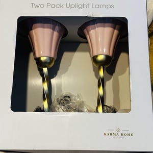May include: A two-pack of uplight lamps in a white retail box labeled Two Pack Uplight Lamps and Karma Home Collection. Each lamp features a fluted, light pink shade atop a metallic gold-toned, twisted stem base, with clear plastic packaging visible.