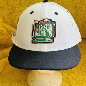 May include: White baseball cap with a black brim. The cap features a patch with the text "BOSTON ALL-STAR GAME '99" in green, red, and white. The cap has a structured fit and a flat brim.