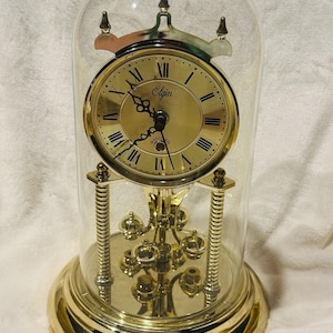 May include: A gold-toned Elgin clock with a glass dome. The clock face is gold with black Roman numerals and hands. The clock has a pendulum and decorative accents. The base and frame are also gold.
