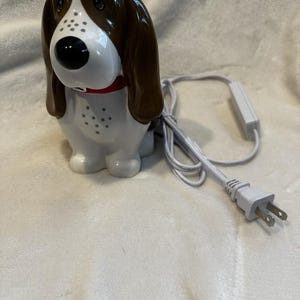 Op de afbeelding: Een glanzende keramische lamp in de vorm van een hond met een wit lichaam, bruine oren en een zwarte neus. De hond draagt een rode halsband en heeft een reeks kleine gaatjes op de borst. Een wit elektrisch snoer met stekker en schakelaar is bevestigd.