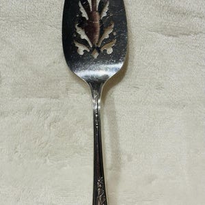 Puede incluir: Cuchara de servir plateada con una cabeza ovalada ranurada. La cabeza presenta un diseño calado decorativo. El mango es delgado, con detalles florales en relieve. Un utensilio vintage para servir comida.