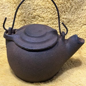 May include: A small, dark brown cast iron teapot with a curved handle and a lid. The teapot has a spout and a textured surface. The handle is made of a dark metal. The teapot is sitting on a yellow surface.