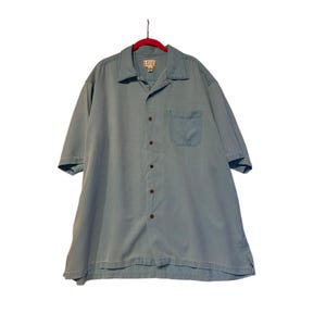 Puede incluir: Una camisa azul claro de manga corta con un bolsillo en el pecho. La camisa tiene cuello y está colgada de una percha roja. La camisa está hecha de una tela suave y ligera.