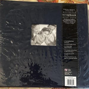 May include: A dark blue post-bound scrapbook measuring 12 inches by 12 inches. The cover features a central square window displaying a black and white photograph of a family. The packaging includes text detailing 10 top-loading sheet protectors.