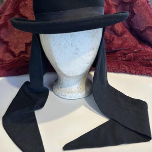 May include: A black hat with a rounded crown and a wide brim. The hat has a black ribbon around the base of the crown and long, black fabric streamers attached to the sides. The hat is displayed on a mannequin head.