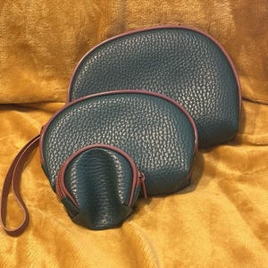 Peut inclure: Un ensemble de trois pochettes semi-circulaires d'un bleu sarcelle foncé avec une texture grainée. Chaque pochette présente une bordure de fermeture éclair contrastante de couleur mauve le long du bord incurvé. La plus petite pochette inclut une dragonne assortie.