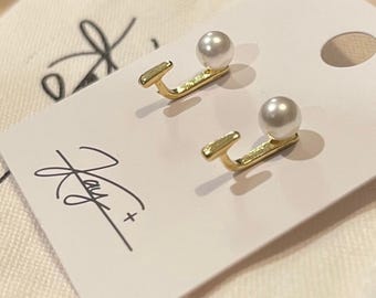 Könnte beinhalten: Ein Paar goldene Ohrringe mit einer kleinen Perle an jedem. Die Ohrringe befinden sich auf einer weißen Karte mit dem aufgedruckten Text "Jay +".