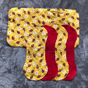 May include: Set of four yellow and red fabric burp cloths with a ladybug and daisy pattern. The cloths are quilted and have a curved shape.