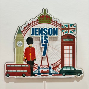Op de afbeelding: Een rode, witte en blauwe taarttopper met een Londens thema. Het heeft een rode dubbeldekkerbus, een rode telefooncel, een zwarte taxi, het London Eye, Big Ben en een koninklijke wacht. De topper zegt "Jenson is 7".