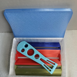 May include: A clear plastic container with a blue lid holds several multi-colored soap-making tools. A light blue tool with holes and a handle sits atop red, blue, and green soap molds. The text "Soap Savvy Products" is visible.