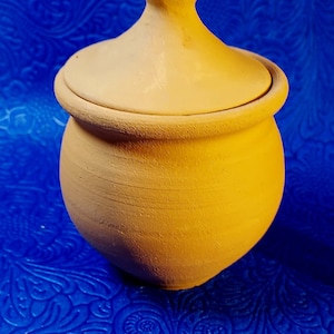 May include: A small, round, terracotta pot with a lid. The pot is a light brown color and has a smooth, slightly textured surface.