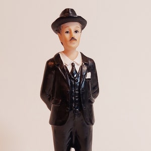 Puede incluir: Una figurita en blanco y negro de un hombre con un traje negro, camisa blanca y corbata negra. Lleva un sombrero fedora negro y tiene bigote. La figurita está de pie sobre una base redonda con una superficie texturizada.