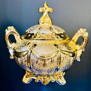 Puede incluir: Un azucarero dorado con tapa, dos asas ornamentadas y cuatro patas. La superficie presenta un acabado mate texturizado con patrones florales en relieve y detalles en oro pulido brillante en el borde, las asas, la base y el remate.