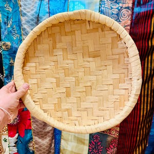 May include: A round, woven bamboo tray with a light tan color. The tray features a tightly woven pattern and a rounded rim. The background includes colorful fabric with floral and geometric patterns.