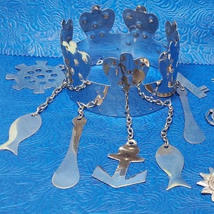 Peut inclure: Une couronne argentée avec un motif en forme de cœur et des breloques pendantes, dont un poisson, une ancre, une cuillère et une clé. La couronne est en métal et a une finition brillante.