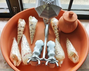 May include: A terracotta dish with a silver umbrella-shaped sculpture in the centre. The sculpture has a chain hanging from it and is surrounded by several seashells. There is also a small terracotta pot with a lid in the dish.