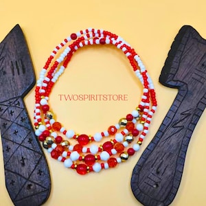 May include: A collection of beaded waist beads in red, white, and gold. The beads are arranged in multiple strands, with the text "TWOSPIRITSTORE" in the center. Two wooden objects flank the beads on a yellow background.