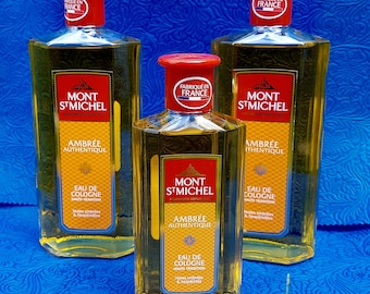 May include: Three bottles of Mont St Michel Eau de Cologne. The bottles are rectangular with red caps and labels. The labels read "Mont St Michel" and "Ambrée Authentique". The bottles are filled with a golden liquid.