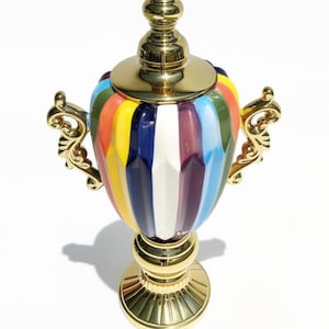 Puede incluir: Una urna decorativa con un cuerpo de cerámica de rayas verticales multicolores con paneles amarillos, azules, blancos y rojo oscuro. La pieza incluye una tapa de metal dorado pulido, una base circular a juego y dos asas de metal dorado ornamentadas.